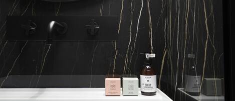 Un baño con pared de mármol negro, un espejo redondo y un lavabo blanco. Hay dos cajas y una botella de jabón de manos sobre el mostrador.