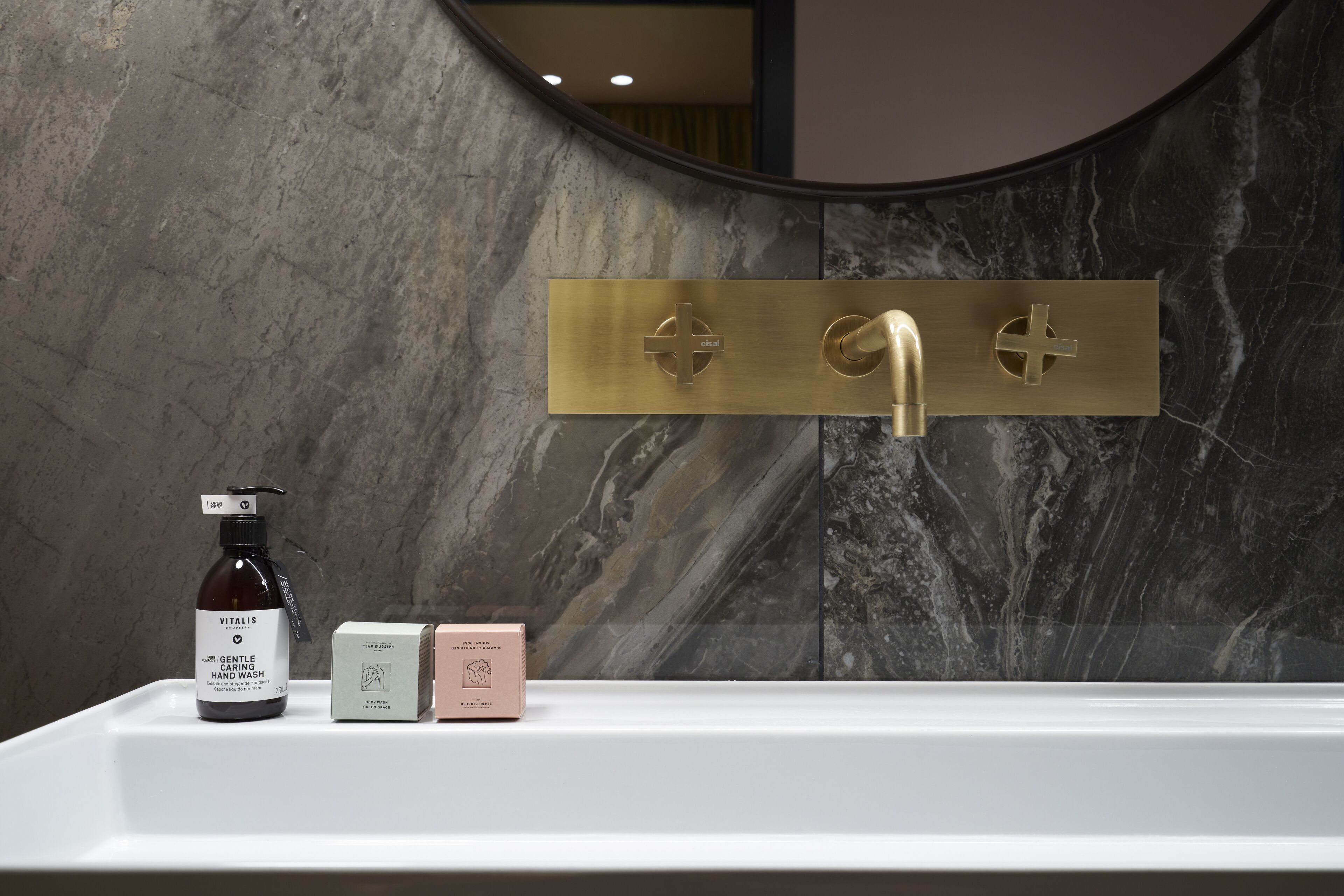 A bathroom with a marble wall, a gold faucet, and a bottle of hand wash soap.