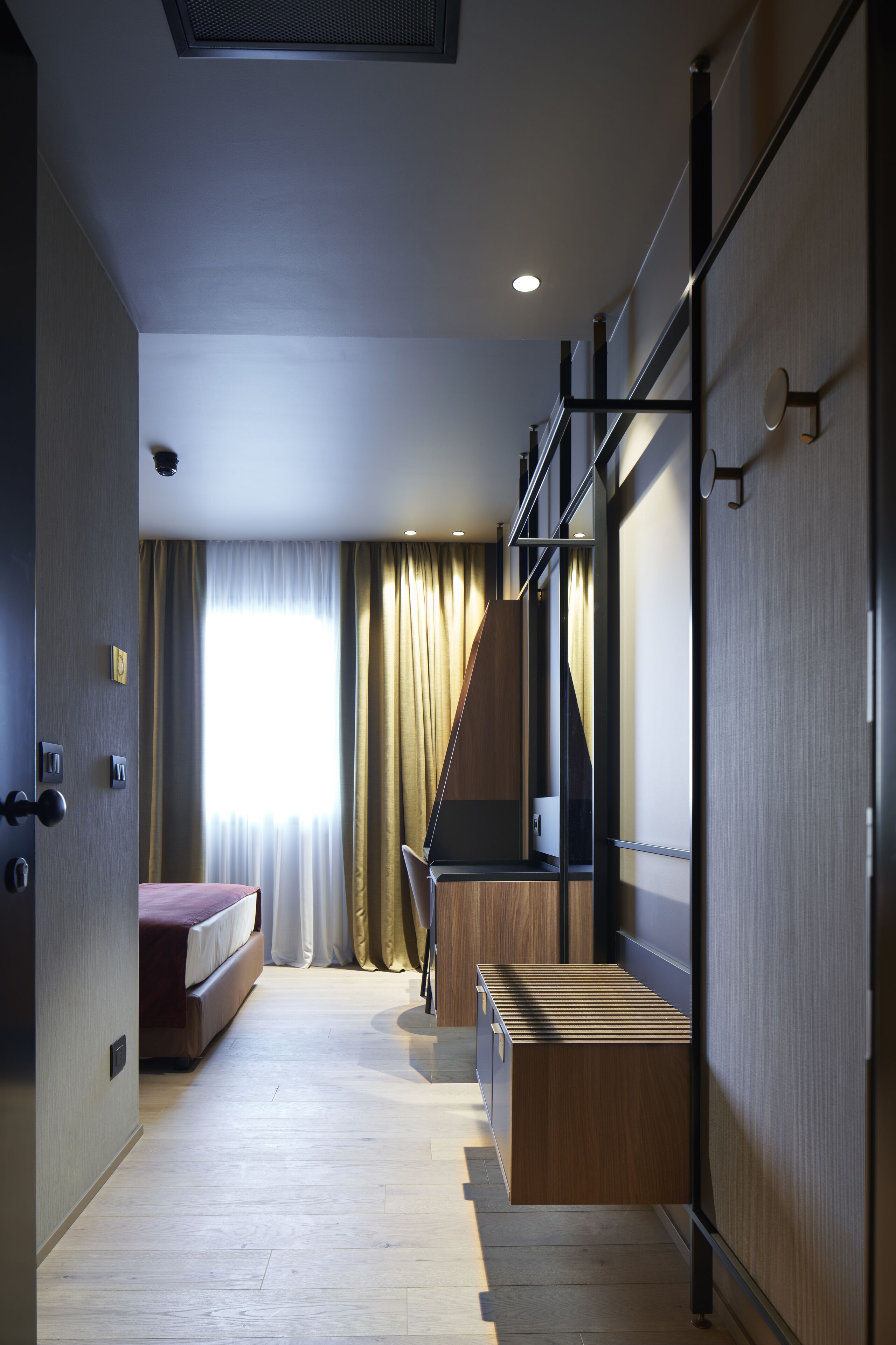 Une chambre d’hôtel moderne, équipée d’un lit, d’un banc en bois, d’un bureau et d’une grande fenêtre avec des rideaux.