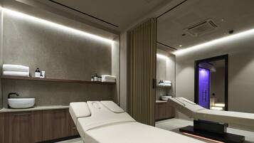 A modern spa room with a large white basin, wooden cabinets, and two reclining massage tables.