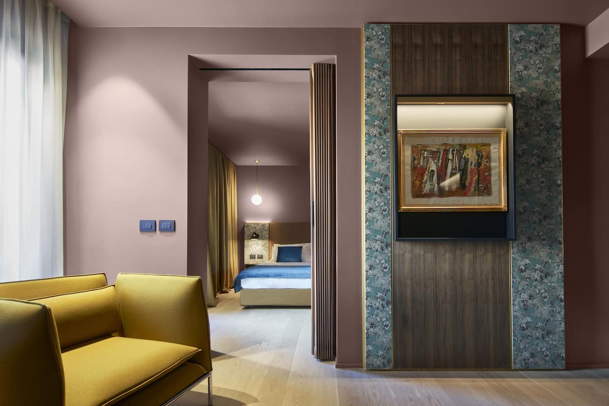 A hotel room with a modern design, featuring a sofa, a bed with a blue headboard, a wall-mounted artwork, and a decorative panel.