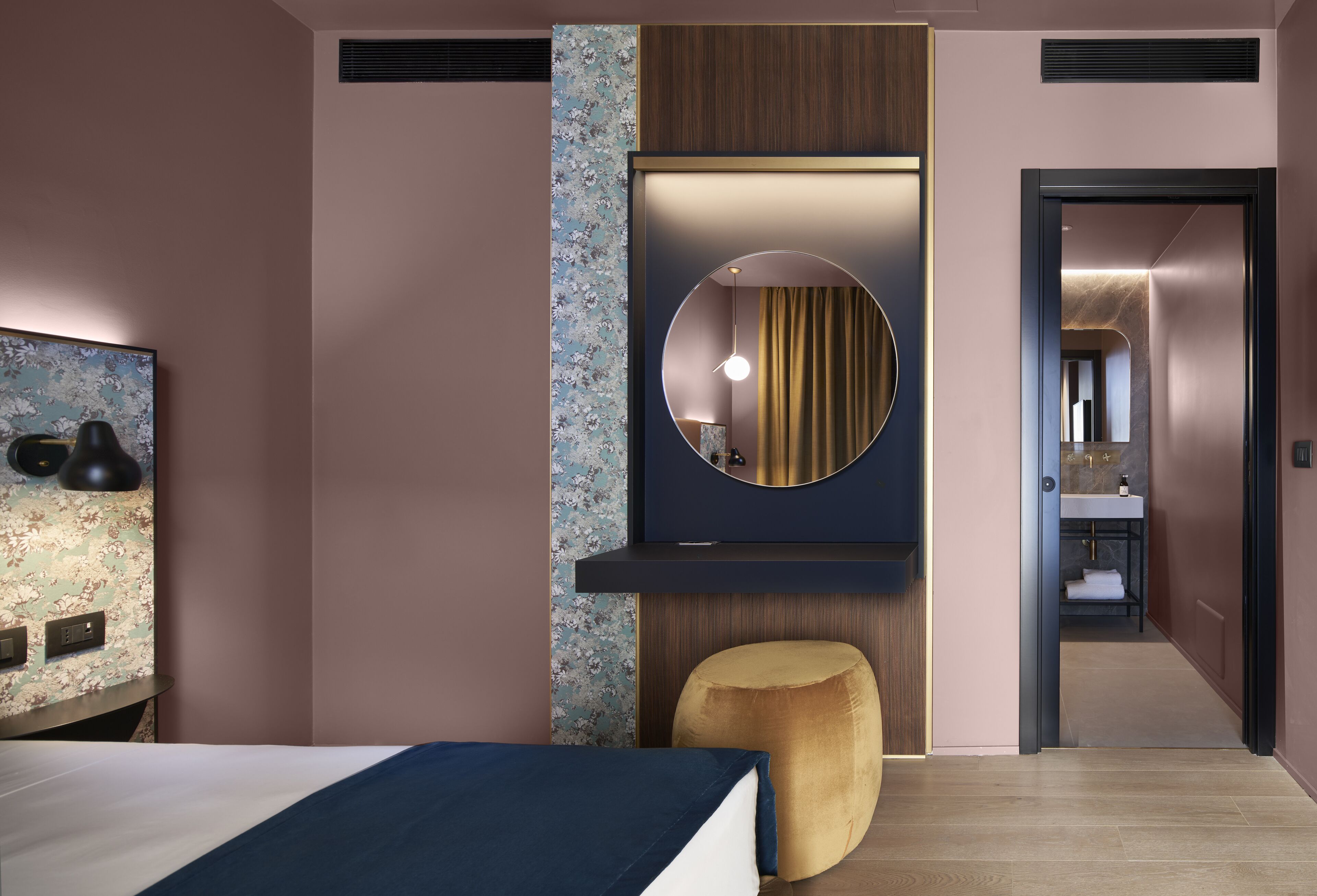 Ein modernes Hotelzimmer mit einem Bett, einem runden Spiegel, einem kleinen Hocker und einem durch eine offene Tür einsehbaren Badezimmer.