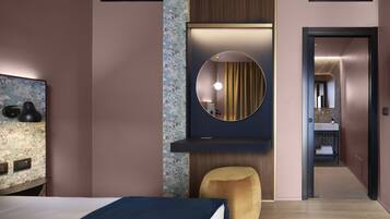 Ein modernes Hotelzimmer mit einem Bett, einem runden Spiegel, einem kleinen Hocker und einem durch eine offene Tür einsehbaren Badezimmer.
