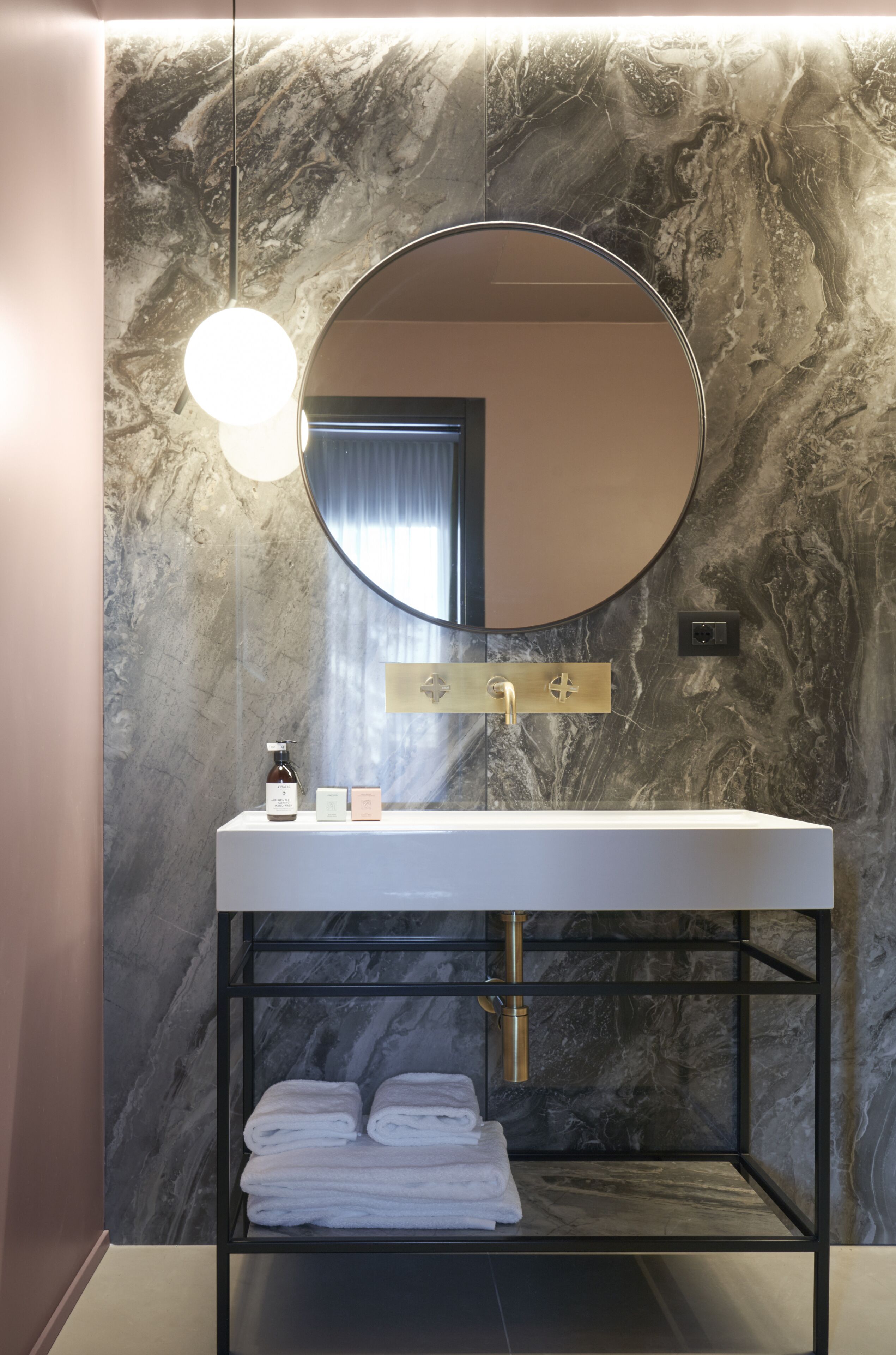 Un baño moderno con pared de mármol, un espejo redondo, un lavamanos blanco y una repisa con toallas dobladas.