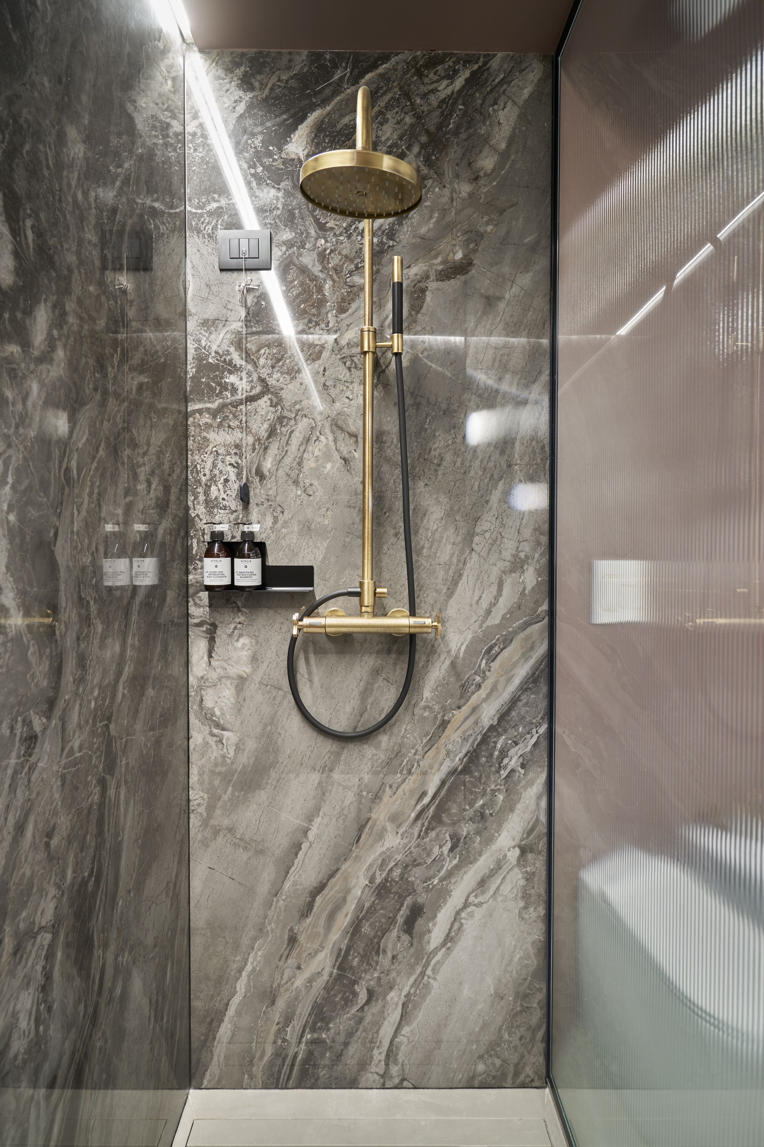 A modern bathroom with a marble wall, a gold showerhead, and a black hose.