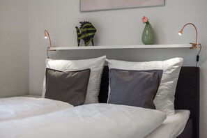 Ein ordentlich bezogenes Bett mit weißen und grauen Kissen, eine grüne Vase mit einer rosa Blüte und ein Regal mit einer Fischdekoration und zwei Lampen.