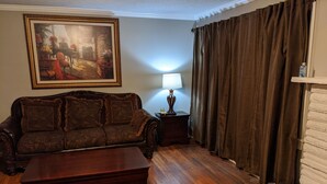 A living room with a dark brown sofa, a wooden coffee table, a painting on the wall, a lamp on a side table, and a curtain. - Cozy lakeview home (Guntersville)