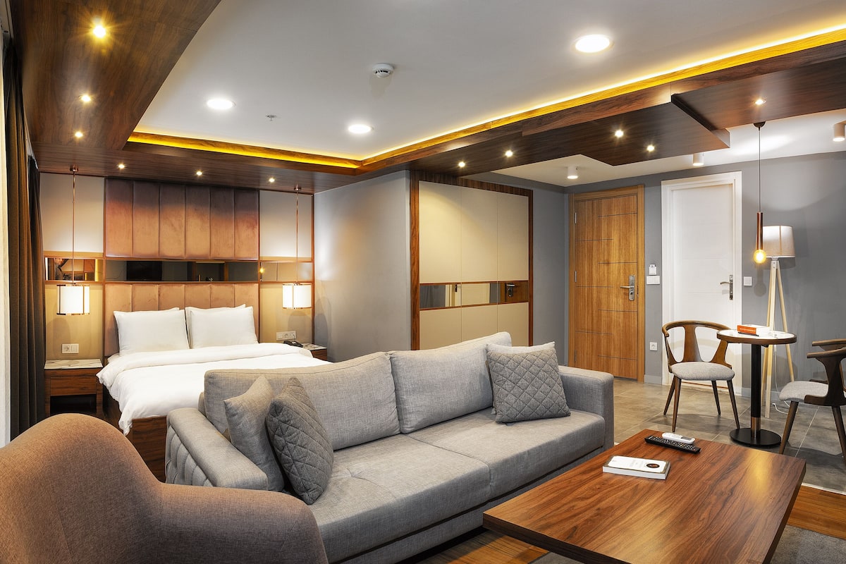 A modern hotel room with a bed, sofa, and armchair, wooden ceiling, and a small table with a remote control.
