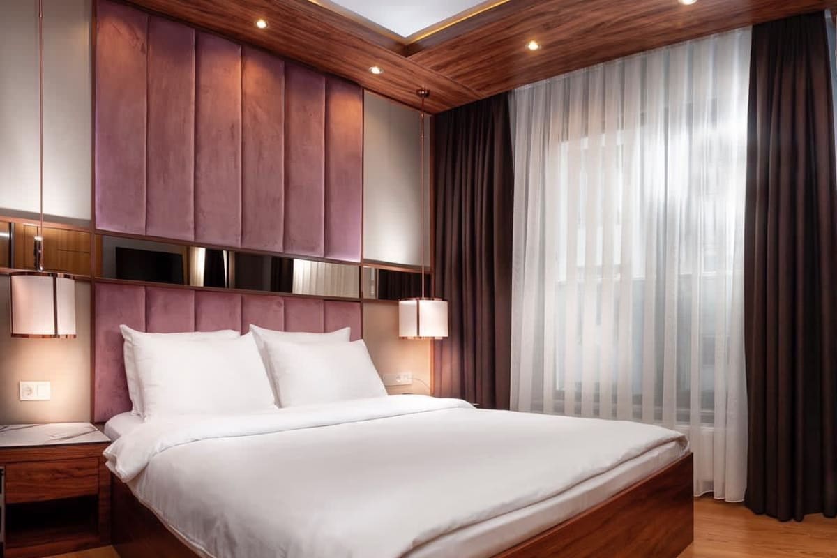 A modern hotel room with a large bed, wooden headboard, and a view of the city through sheer curtains.