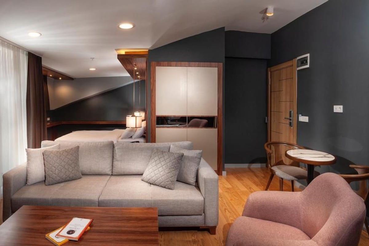A modern living room with a sofa, a coffee table, and a chair. There is a wooden door and a small table with a lamp.
