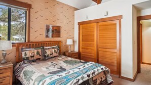 Quarto com cama grande, cabeceira de madeira e mesa de cabeceira com luminária. Há uma janela com vista para árvores e um guarda-roupa de madeira com portas de correr.