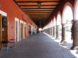 A covered walkway with arches, shops on the left, and a view of a building in the distance. - Hotel Santa Rosa (San Pedro Cholula)