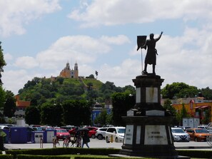 A statue on a pedestal with a plaque, surrounded by a parking lot with cars and a building in the background. - Hotel Santa Rosa (San Pedro Cholula)