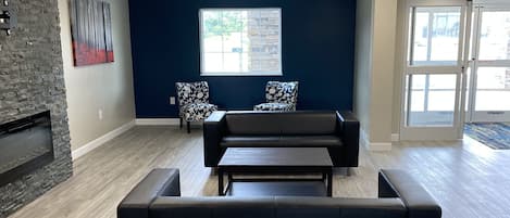 A spacious lobby with a dark blue wall, a stone fireplace, two patterned chairs, a black sofa, and a wooden floor.