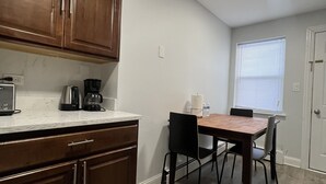 Una cocina con armarios de madera, una mesa de comedor con sillas, una cafetera y una ventana con persianas.