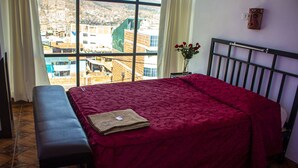 Una habitación con cama, un banco, una ventana con vista a la ciudad y un florero con flores.