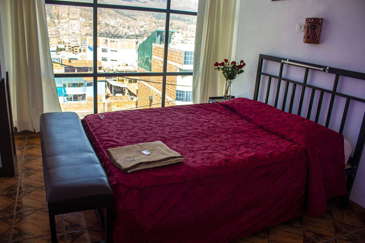 A room with a bed, a bench, a window with a city view, and a vase with flowers.