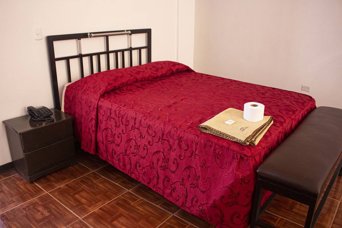 A neatly made bed with a red patterned bedspread, a bedside table with a phone, and a bench.