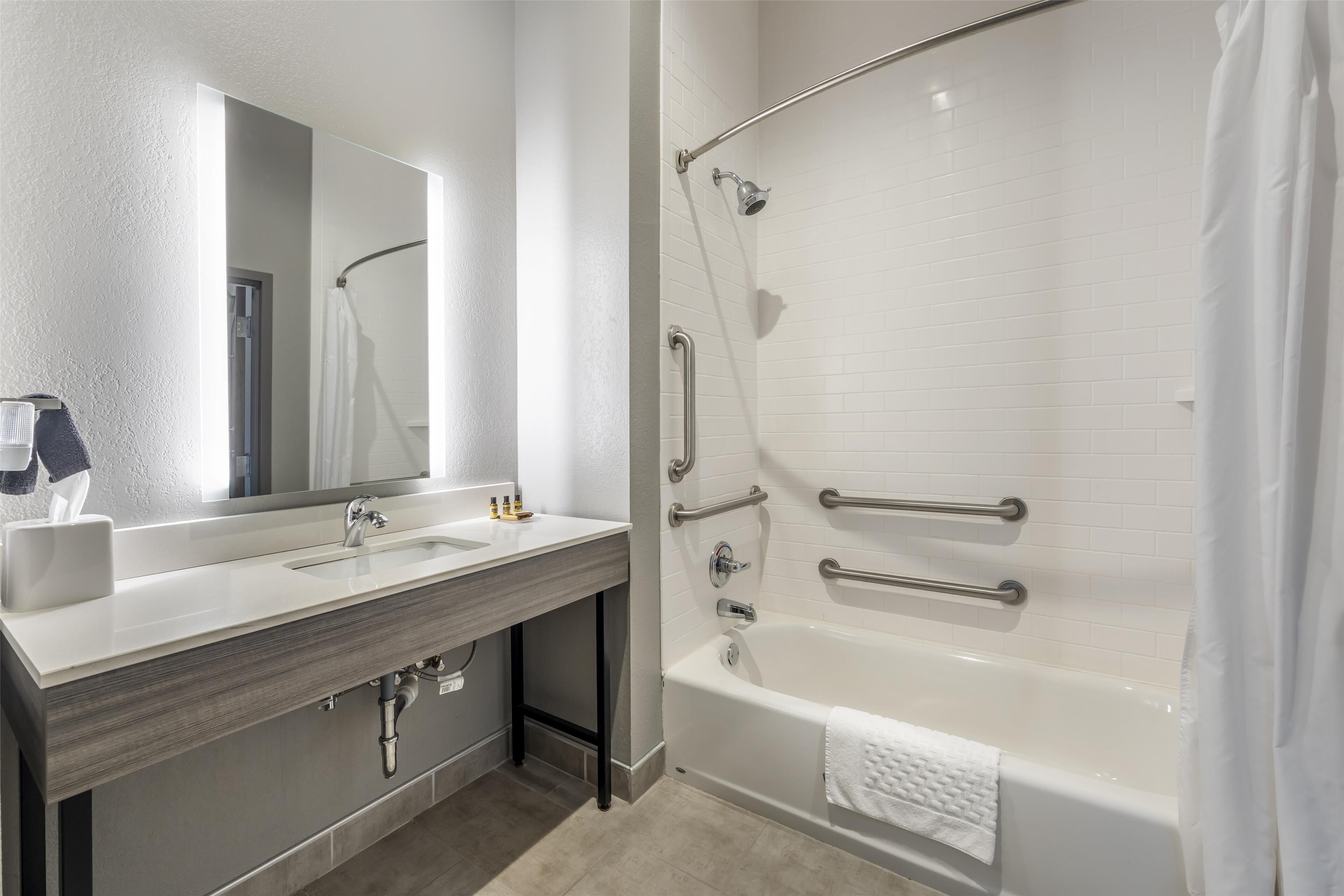 A modern bathroom with a white sink, mirror, and bathtub. It includes a shower with a grab bar and a towel rack.