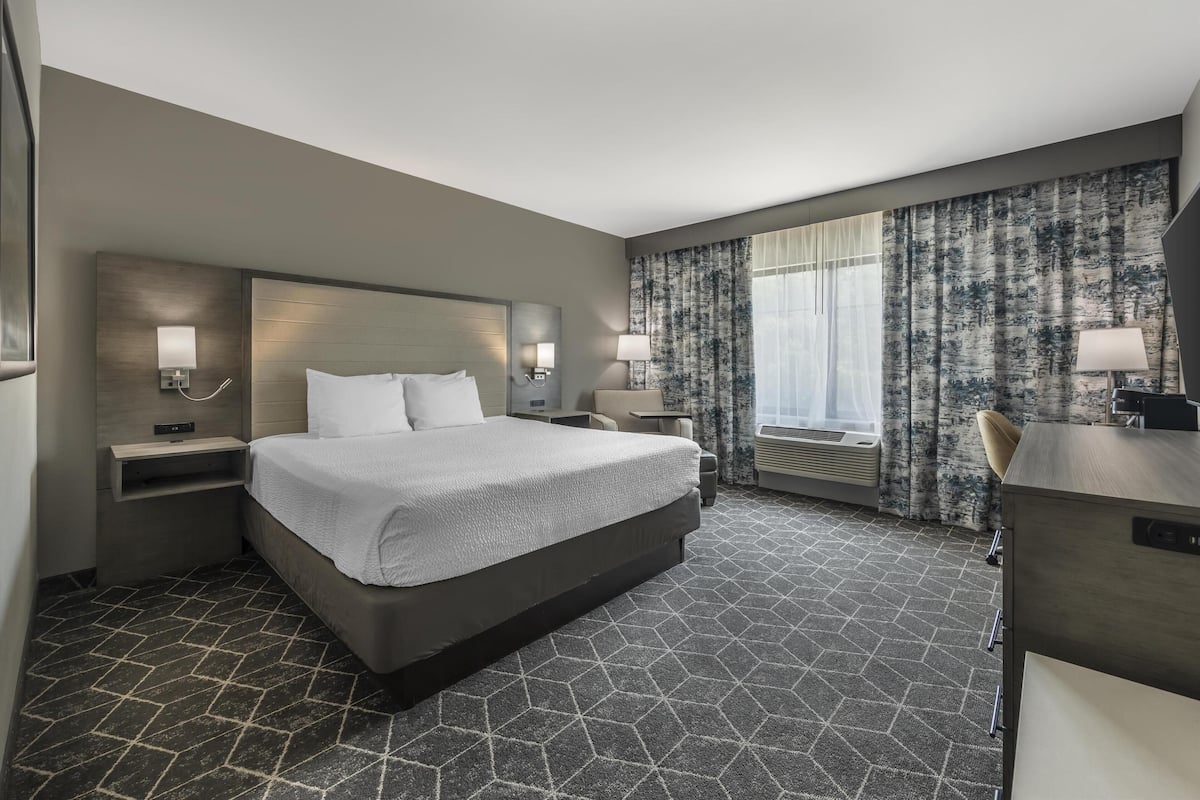 A hotel room with a large bed, two bedside tables with lamps, a desk with a chair, and a large window with curtains.