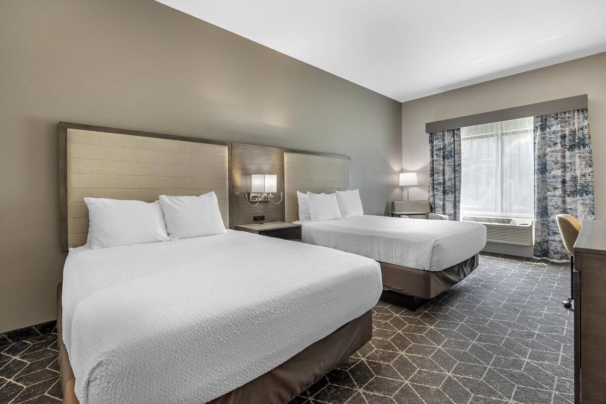 A hotel room with two beds, a large window with curtains, a nightstand with a lamp, and a wall-mounted mirror.