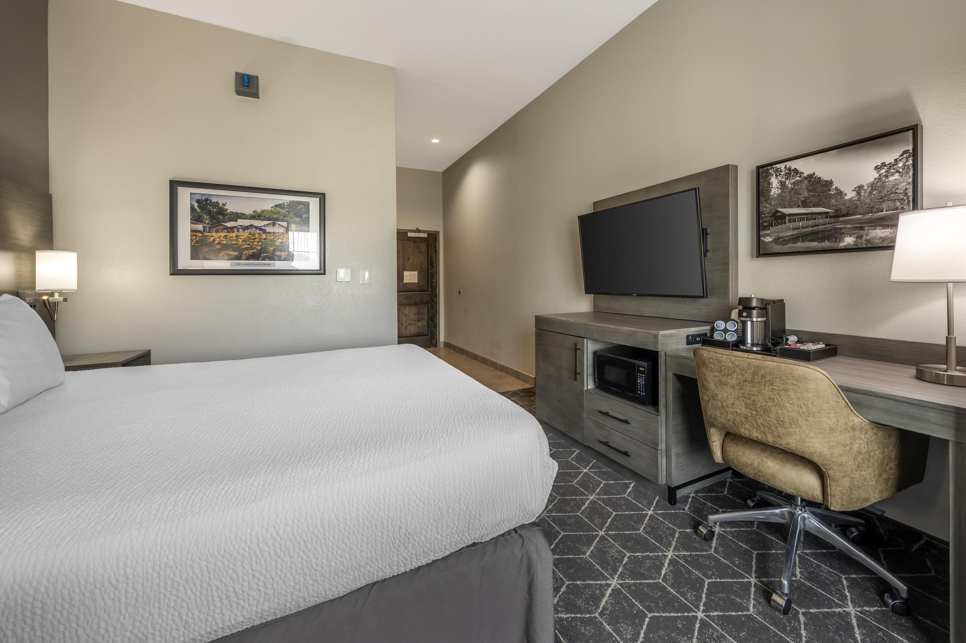 A hotel room with a bed, a desk with a chair, a flat-screen TV, and artwork on the walls.