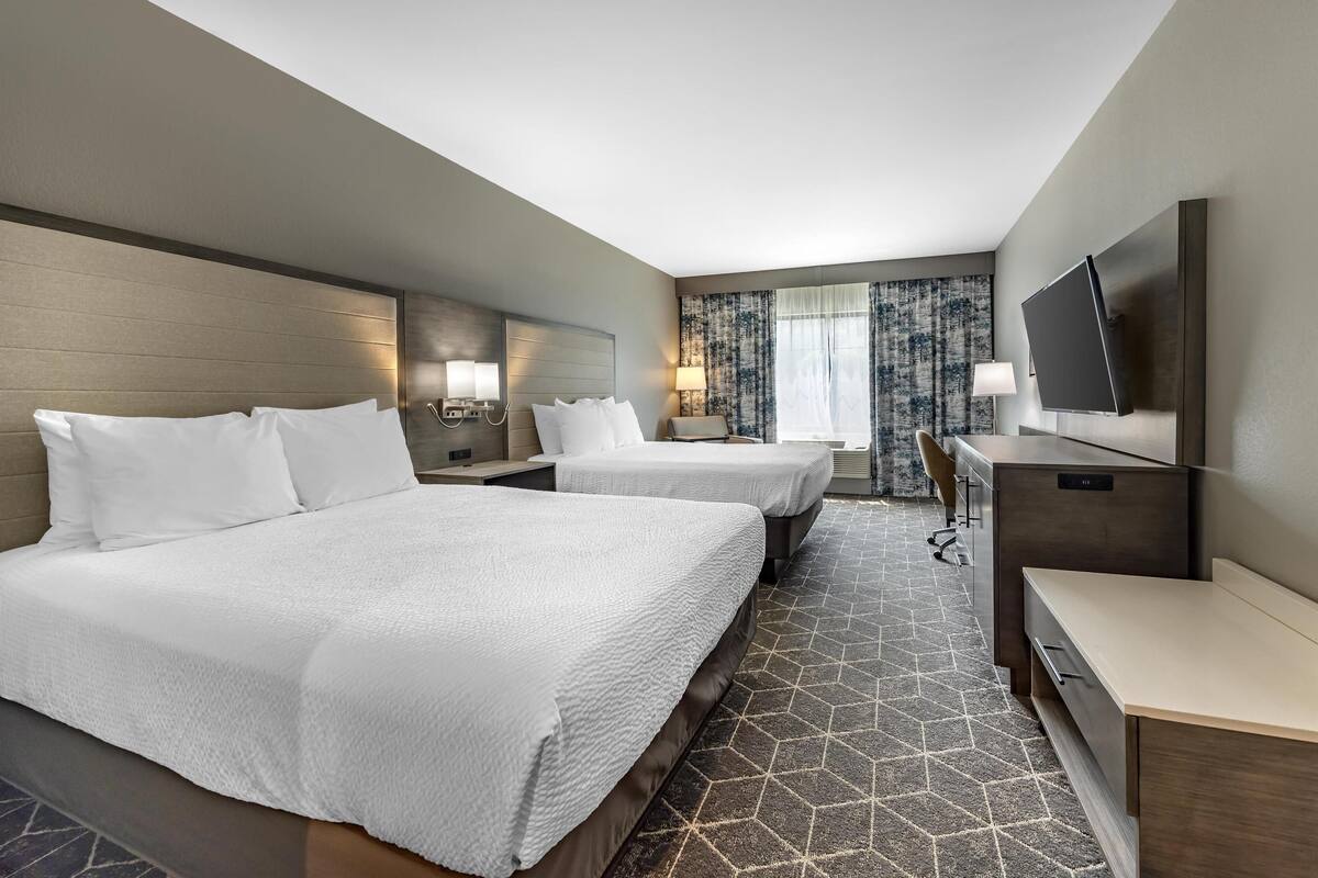 A hotel room with two beds, a desk, a TV, and a large window with curtains.