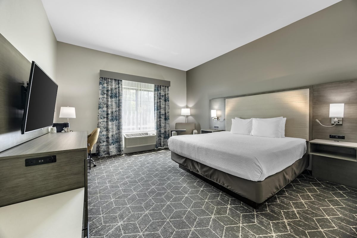 A hotel room with a large bed, two bedside tables with lamps, a flat-screen TV, a desk with a chair, and a window with curtains.