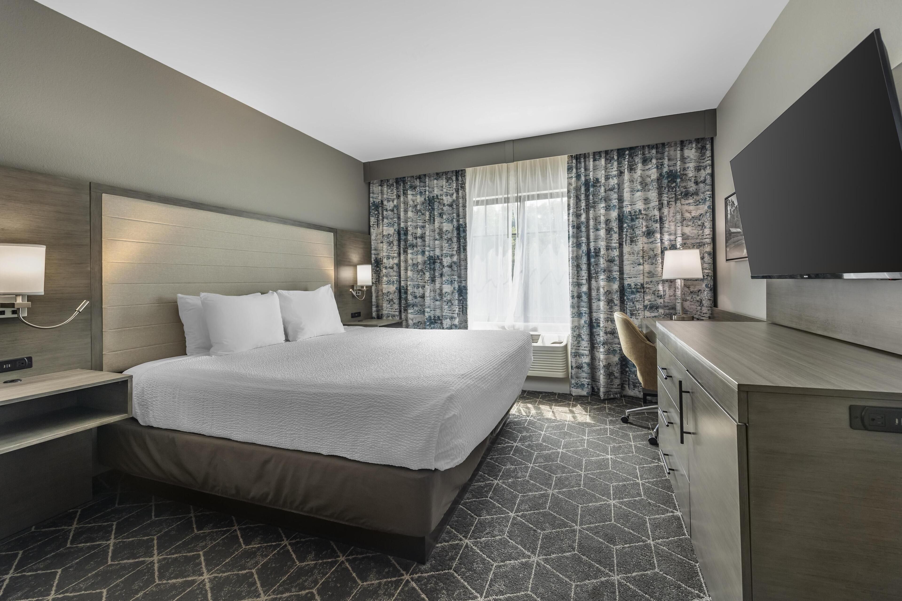 A modern hotel room with a large bed, a desk with a flat-screen TV, and a window with patterned curtains.