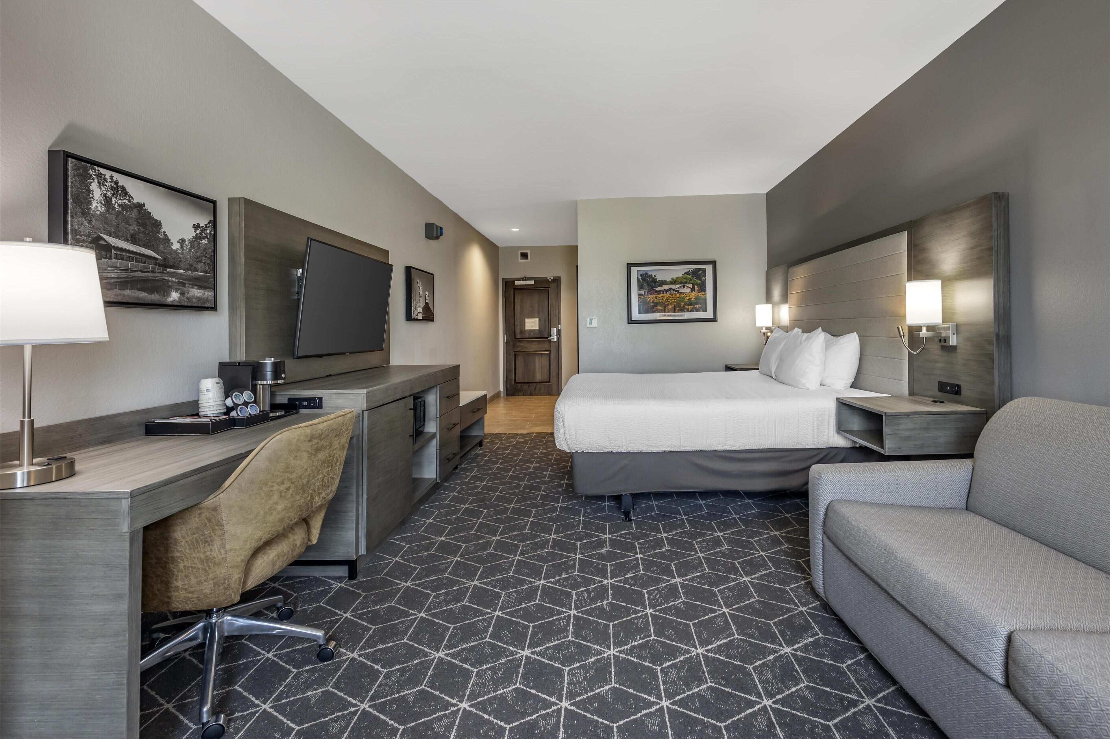 A hotel room with a bed, a sofa, a desk with a chair, a TV, and a lamp.