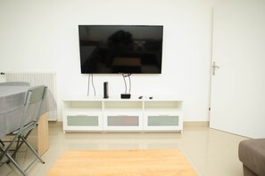 Uma sala de estar com uma televisão de tela plana fixada em um móvel branco, uma mesa de jantar com toalha de mesa cinza e um sofá marrom.