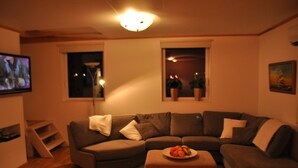 A living room with a brown sofa, a small table with fruit, a TV, and two windows with plants.
