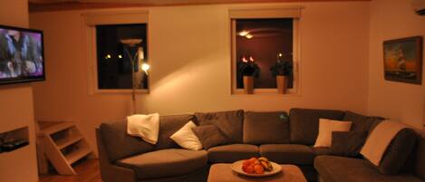A living room with a brown sofa, a small table with fruit, a TV, and two windows with plants.