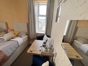 A hotel room with a bed, a small table with a tray of items, a chair, and a view of the city through the window. - Harbour Lodge B&B (Paignton)