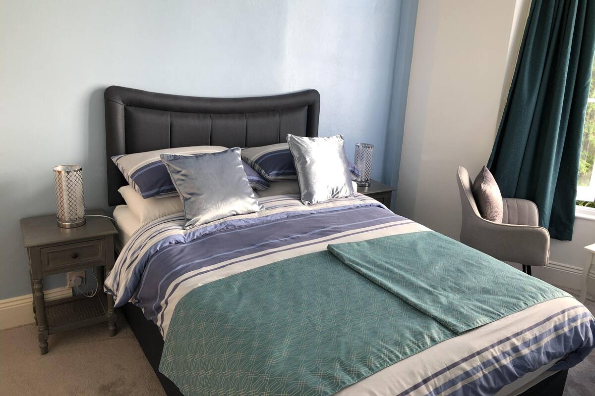 A neatly made bed with a striped bedspread and pillows, a bedside lamp, and a chair by the window.