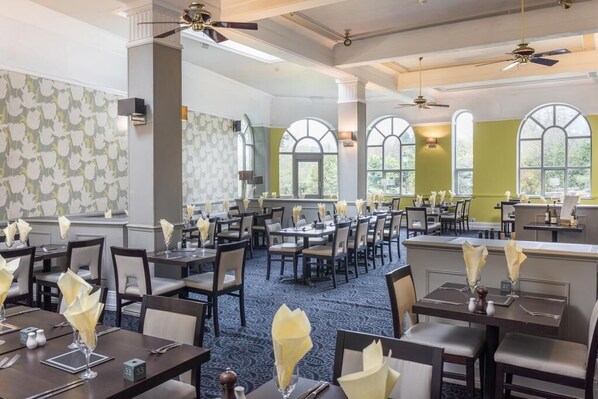 A hotel dining area with multiple tables and chairs, large windows, and ceiling fans.
