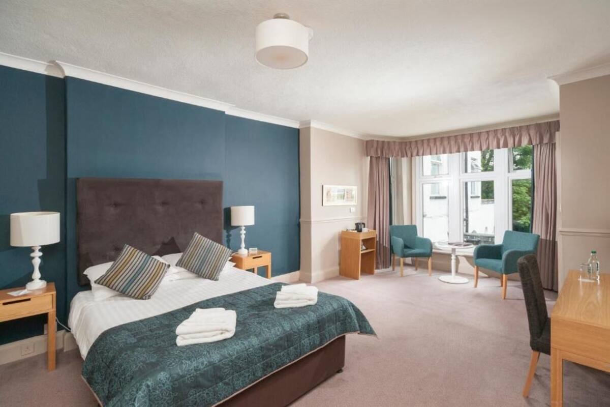 A hotel room with a large bed, bedside tables with lamps, a desk with chairs, and a window with curtains.