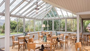 A conservatory with multiple tables and chairs, large windows, and a chandelier. - The Derwentwater Hotel (Keswick)
