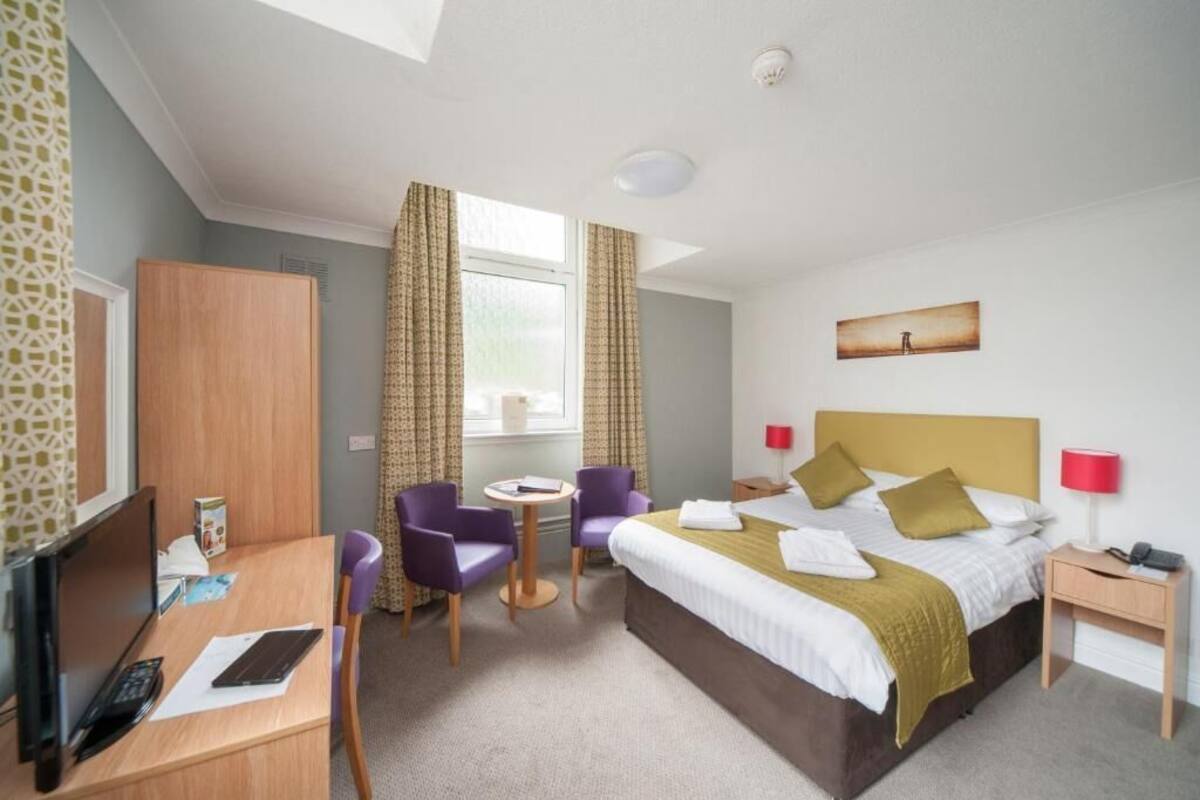 A hotel room with a bed, desk, chairs, a TV, and a window with curtains.