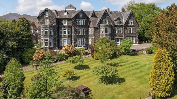 A large, historic stone building with multiple gabled roofs and a well-maintained lawn in the foreground. - The Derwentwater Hotel (Keswick)