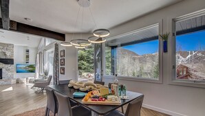 Un comedor moderno con una mesa de madera dispuesta para una comida, ventanas amplias que ofrecen vistas al exterior y una chimenea de piedra al fondo.