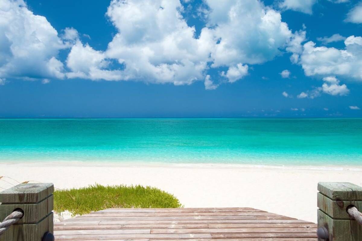 Eine Holzterrasse führt zu einem Strand mit klarem türkisfarbenem Wasser und einem weißen Sandstrand unter blauem Himmel mit Wolken.