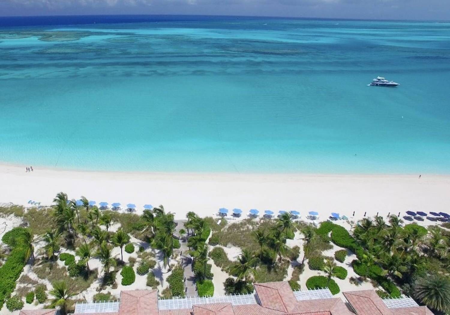 Ein Strand mit klarem türkisblauem Wasser, ein Boot in der Ferne sowie Reihen von Strandsonnenschirmen und Palmen.
