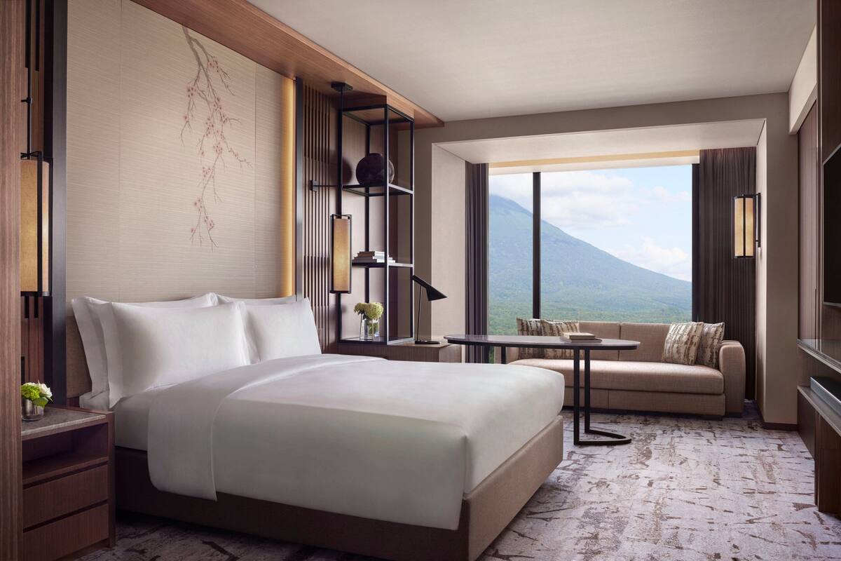 A hotel room with a large bed, a sofa, a small table, a TV, a bookshelf, and a view of a mountain.