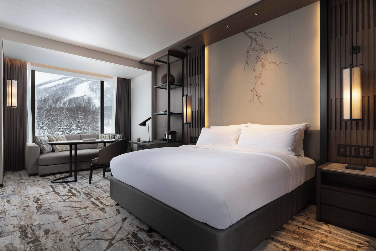 A modern hotel room with a large bed, a seating area with a table, and a view of snow-covered trees.