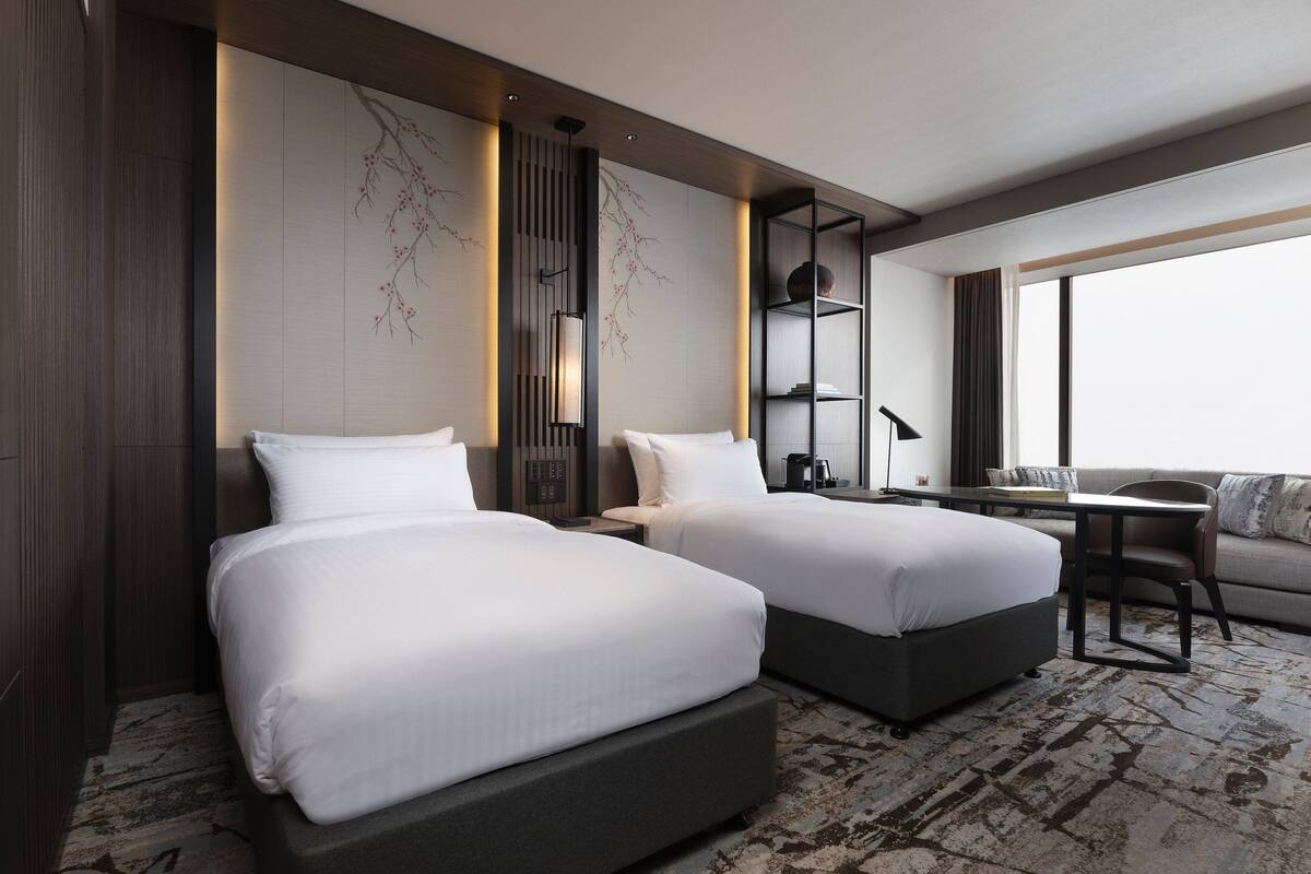 A modern hotel room with two beds, a desk, a sofa, and a large window.