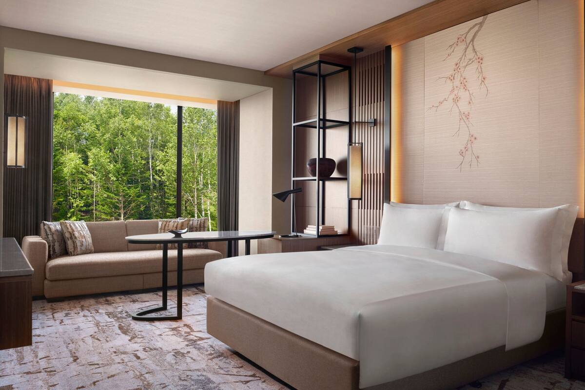 A hotel room with a large bed, a sofa, a small table, and a bookshelf. There is a large window with a view of trees.