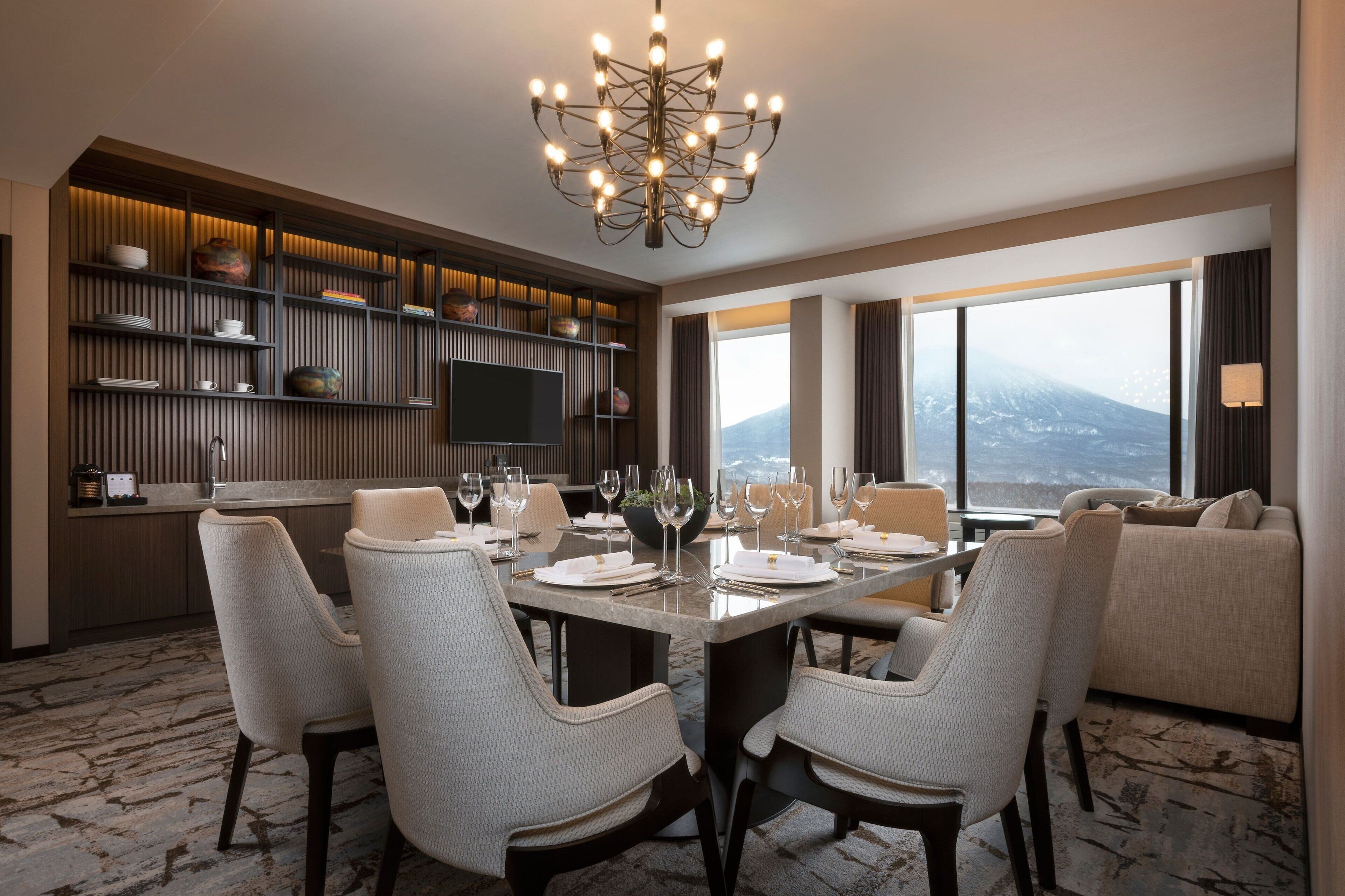 A modern dining area with a large table set for four, surrounded by upholstered chairs. A television is mounted on the wall, and there are shelves with decorative items above. The room has a large window with a view of a mountain range.