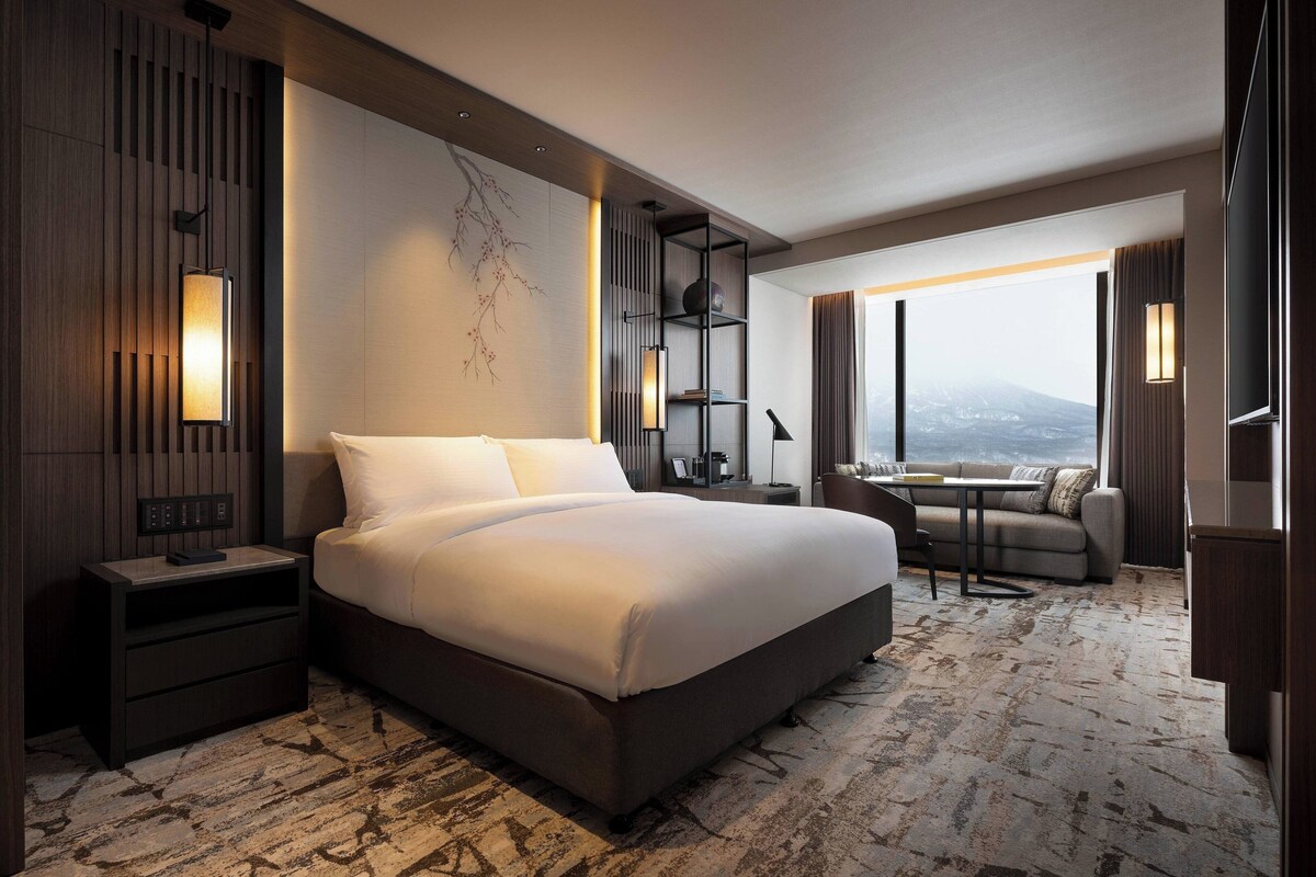 A modern hotel room with a large bed, a desk, and a view of a cityscape.