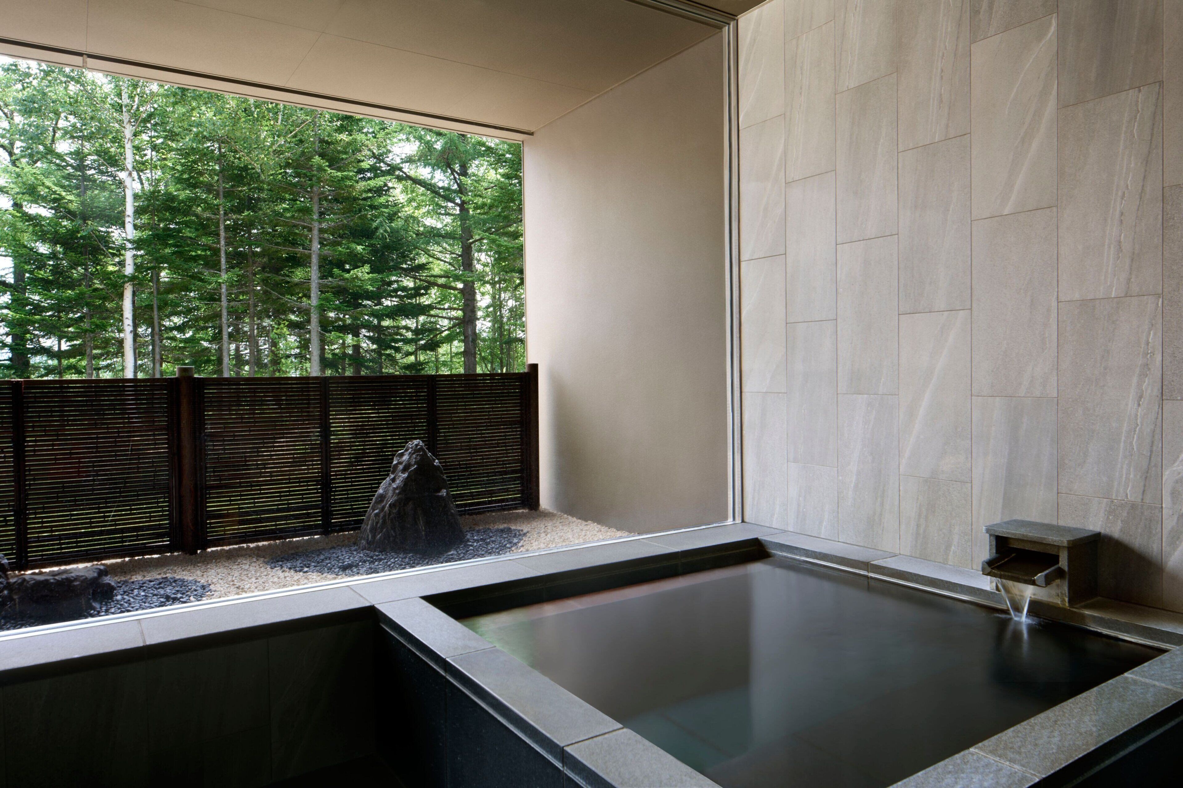 Zona de spa con un jacuzzi, vista a un bosque a través de una ventana y una pared de piedra.